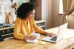 Alt text - A lady working from a kitchen table at home, with her left hand she’s typing on her laptop keyboard and with her right hand she’s writing in a notepad.